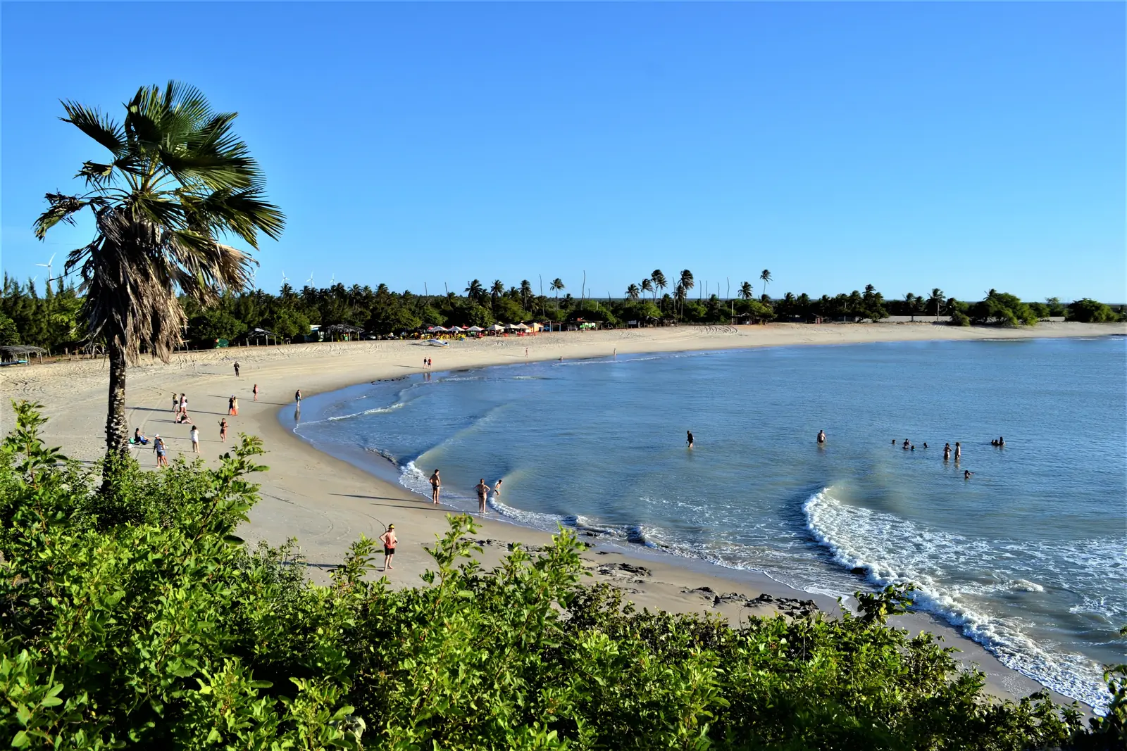 Curved white-sand bay with turquoise water, palm trees and beach tents near São Miguel do Gostoso