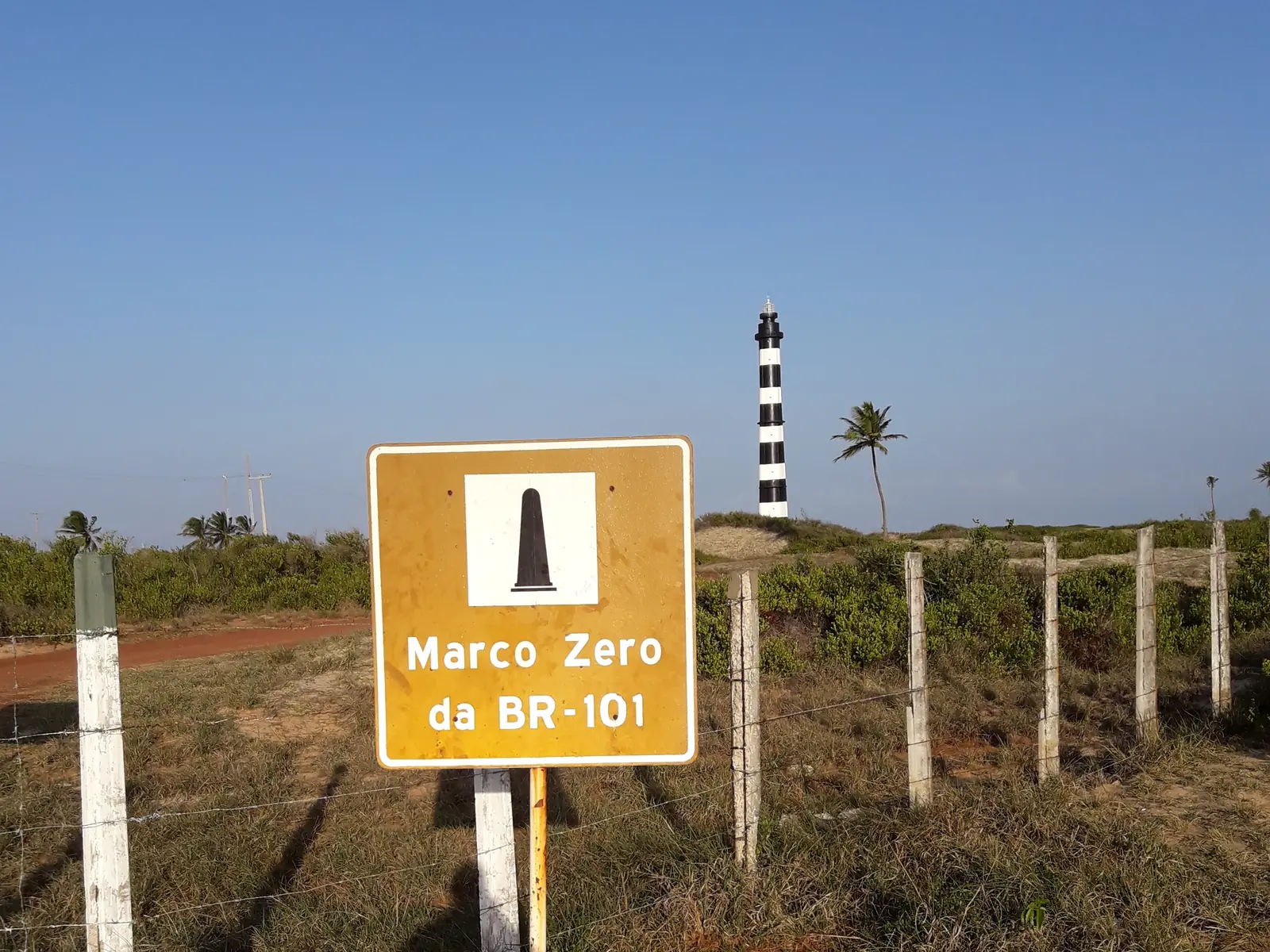 Marco Zero da BR-101 road sign by a sandy track, with the black-and-white Calcanhar lighthouse behind