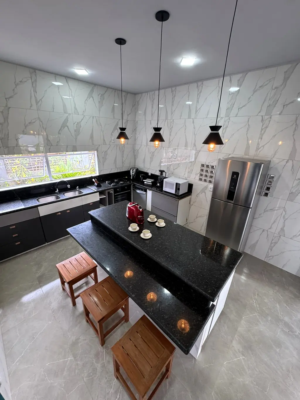 Kitchen with black granite island and two wooden stools, three pendant lamps, steel fridge and marble-look walls