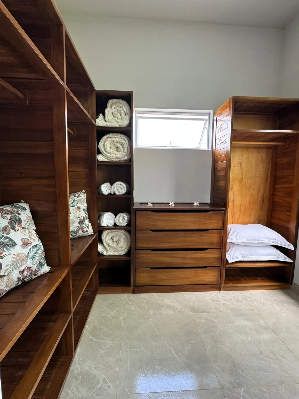 Walk-in wardrobe with open wooden shelves, rolled towels and a cushion, window at the back