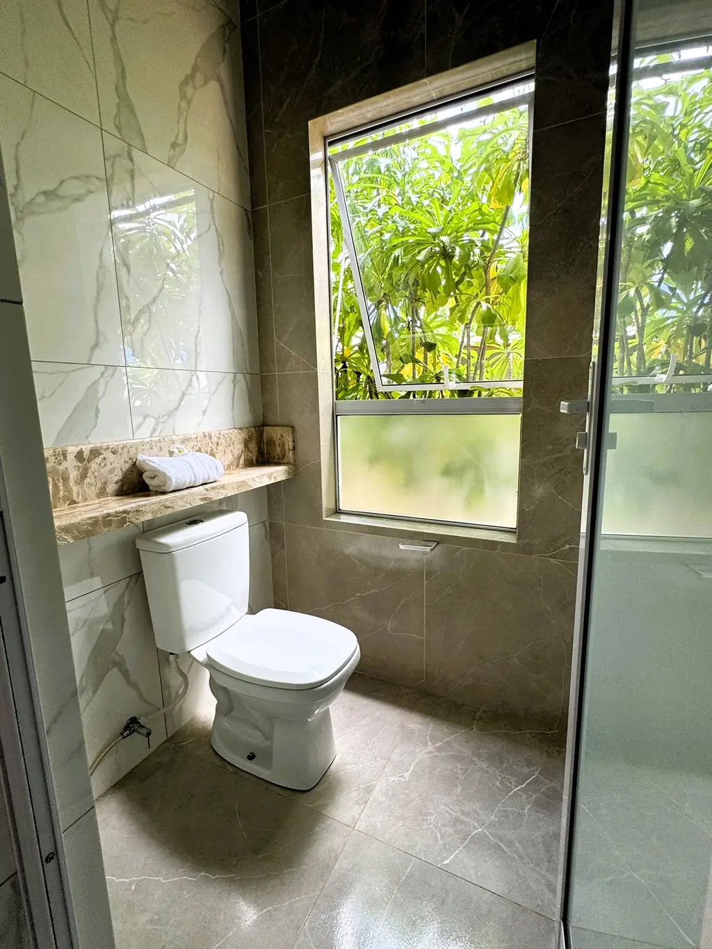 Bathroom with toilet, glass shower screen and a large window onto green foliage