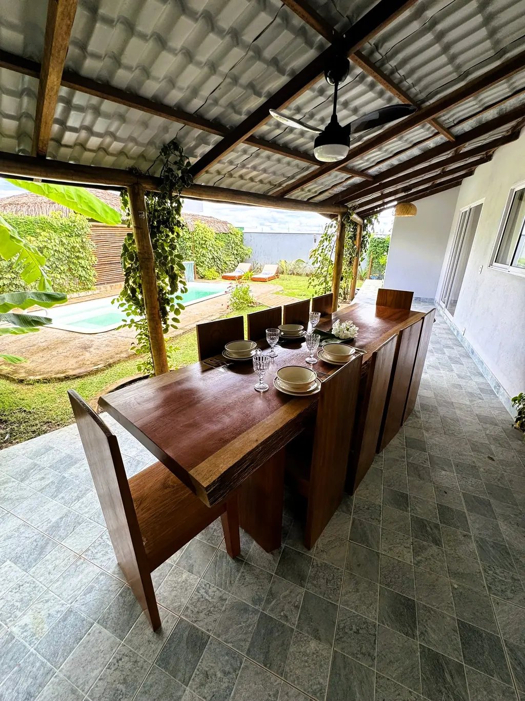 Wooden dining table with chairs under a metal-roofed terrace, garden and pool on the left