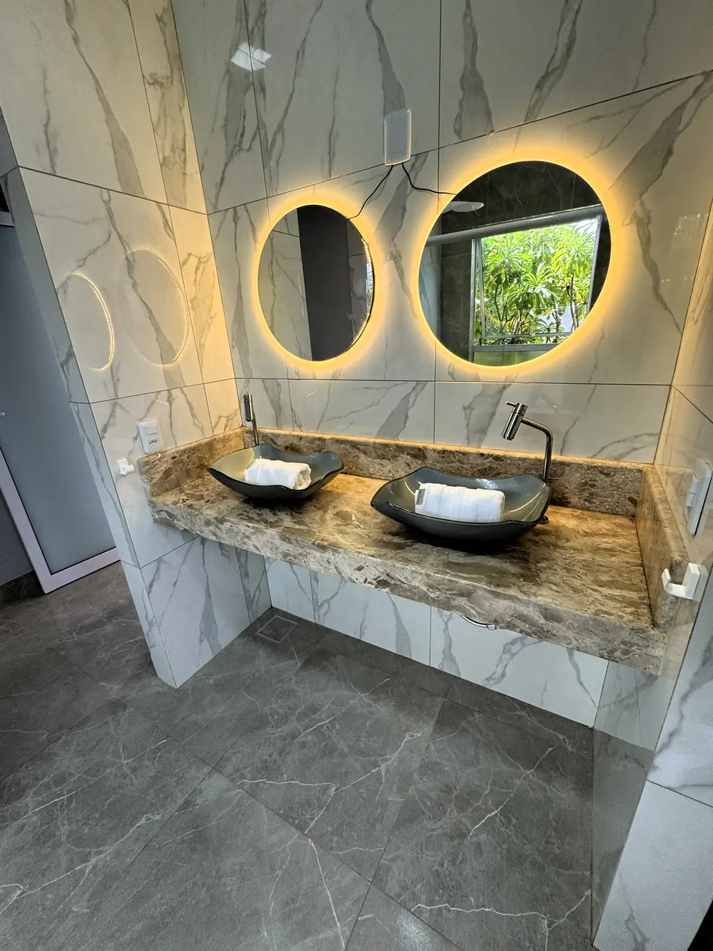 Bathroom with two round backlit mirrors over a stone vanity with twin black basins
